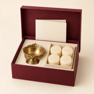 Burgundy gift box holding one brass diya, a pack of ivory candles and a blank cream card